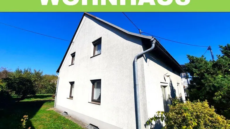 Weißes Einfamilienhaus mit Satteldach und großem Grundstück unter blauem Himmel.
