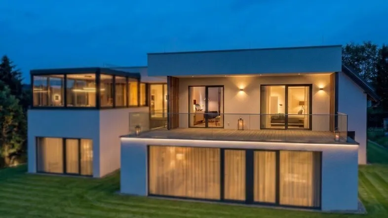 Modernes Haus mit großen Fenstern und beleuchteter Terrasse bei Dämmerung, umgeben von Grün.