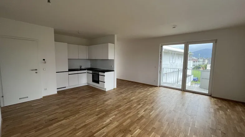 Heller offener Wohnbereich mit integrierter Küche, Parkettboden und Zugang zum Balkon.