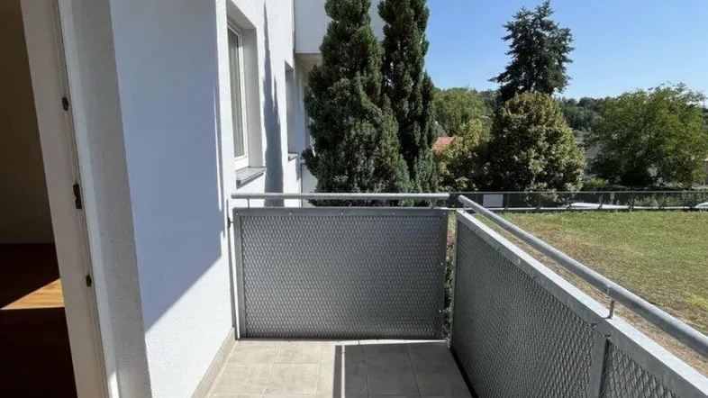 Heller Balkon mit Fliesenboden, Metallgeländer und Blick ins Grüne an einem sonnigen Tag.
