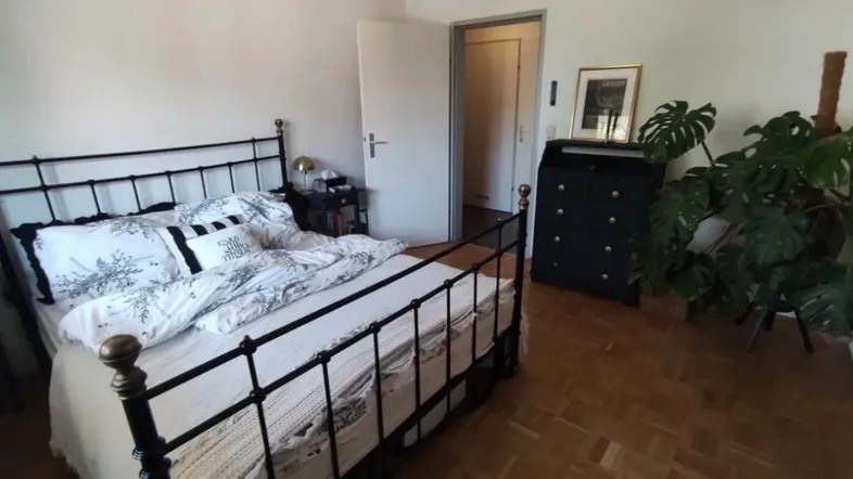 Schlafzimmer mit schmiedeeisernem Bett, dunkler Kommode und großer Grünpflanze auf Parkettboden.