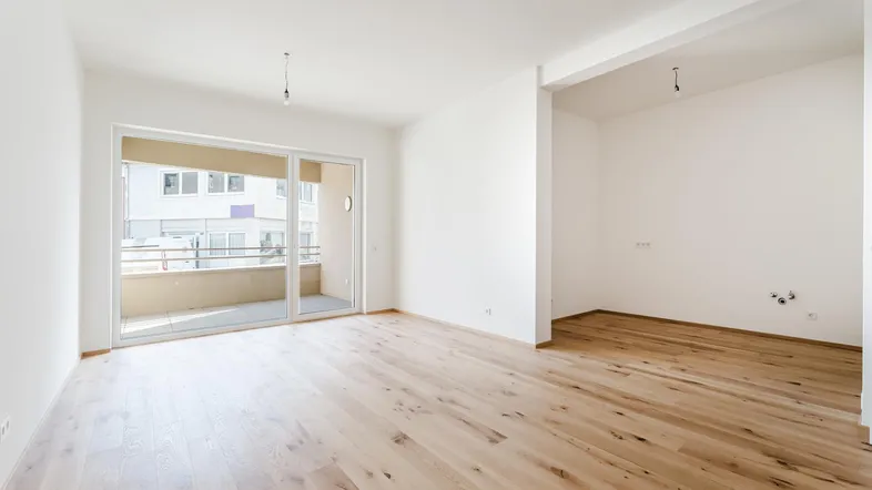 Heller, leerer Wohnbereich mit Holzboden, großen Schiebetüren und Zugang zu einem Balkon.