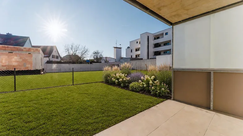Sonnige Terrasse mit angrenzendem Rasen und Blumenbeeten, umgeben von einer Hecke.