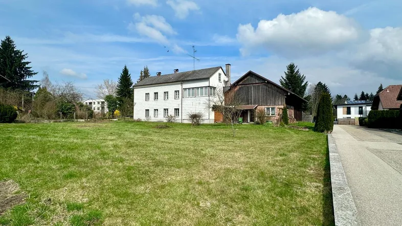 Weitläufige Grünfläche vor einem älteren Wohnhaus mit angrenzendem Scheunengebäude unter blauem Himmel.