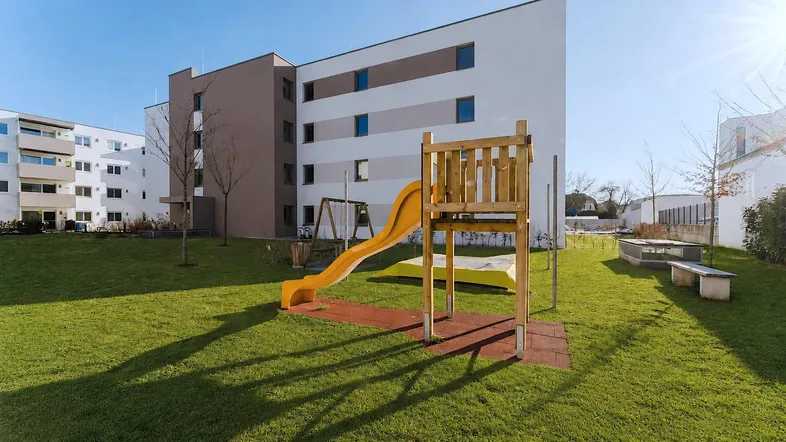 Spielplatz mit Rutsche und Schaukeln auf einer grünen Rasenfläche vor modernen Wohngebäuden.