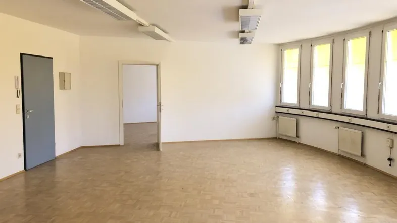 Geräumiges, leeres Büro mit hellem Parkettboden, vielen Fenstern und Zugang zu einem weiteren Raum.