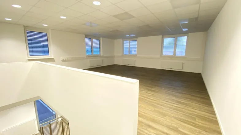 Heller, leerer Raum mit Holzoptikboden, weißen Wänden und mehreren Fenstern, ideal für Büronutzung.