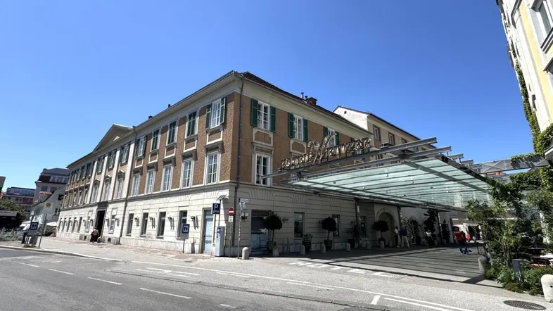 Historisches Gebäude mit klassischer Architektur, grünen Fensterläden und modernem Glaseingangsbereich an einer Straße.
