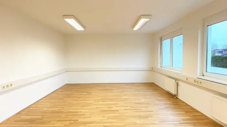 Heller, leerer Büroraum mit Holzfußboden, weißen Wänden und großen Fenstern, die viel Tageslicht hereinlassen.