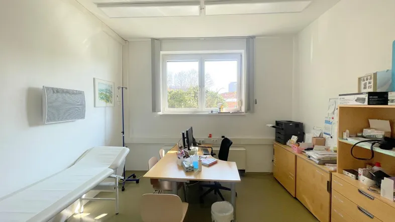 Heller Behandlungsraum mit Untersuchungsliege, Schreibtisch und Stauraum, gut beleuchtet durch ein Fenster.