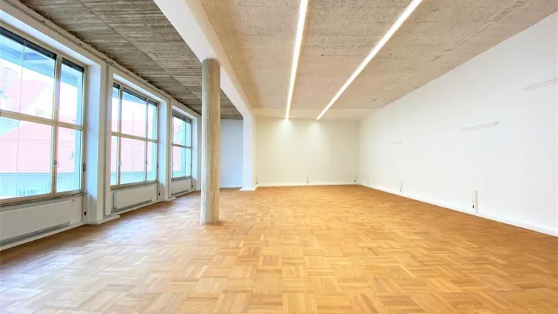Geräumiger, heller Büroraum mit großen Fenstern, Holzboden und einer freiliegenden Betondecke.