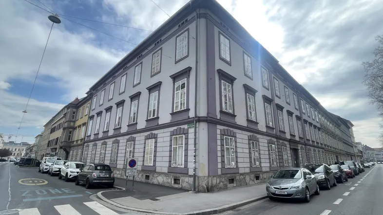 Historisches Gebäude an einer Straßenecke mit vielen Fenstern und parkenden Autos unter bewölktem Himmel.