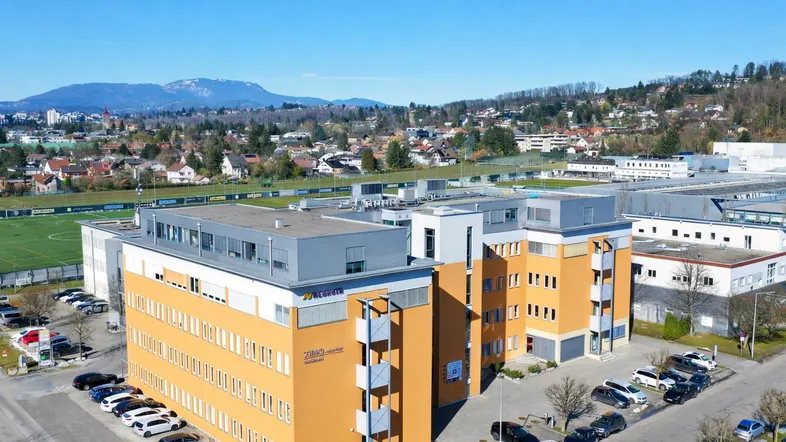 Luftaufnahme eines modernen Bürogebäudes mit orangefarbener Fassade und umliegenden Parkplätzen vor bergiger Kulisse.
