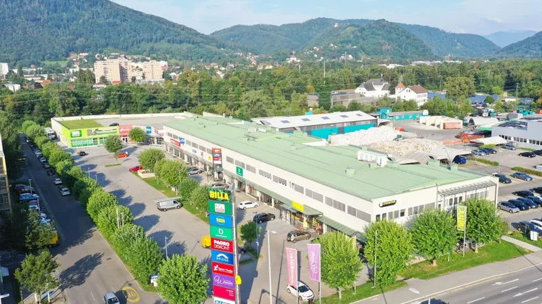 Luftaufnahme eines großen Gewerbekomplexes mit Parkplätzen und umliegender grüner Landschaft und Bergen.