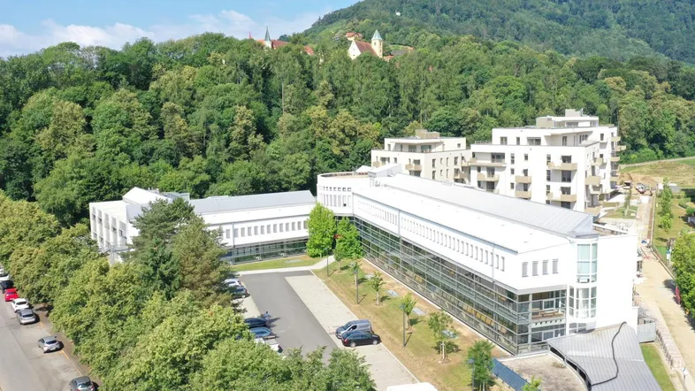 Luftaufnahme eines modernen Bürogebäudes mit umliegender Grünfläche und weiteren Neubauten im Hintergrund.