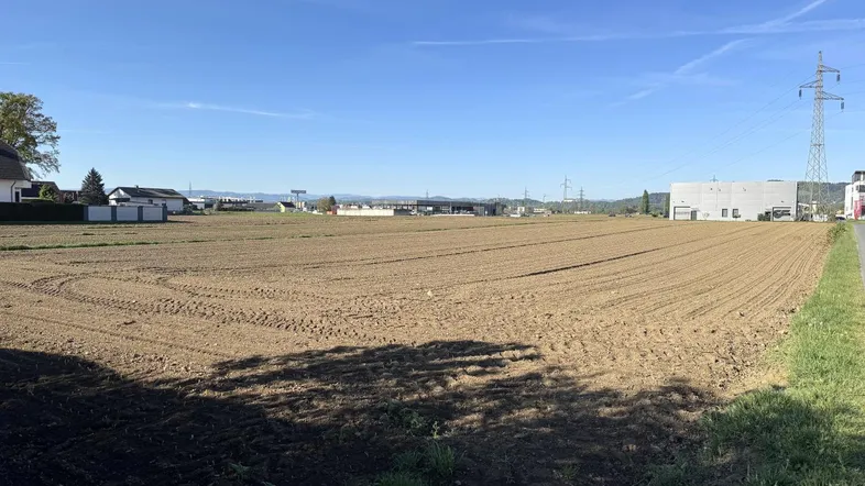 Weites, unbebautes Feld unter blauem Himmel mit angrenzender Bebauung und Strommasten im Hintergrund.