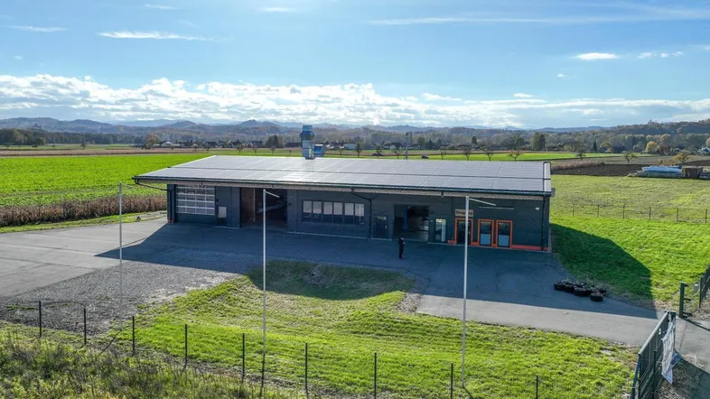 Luftaufnahme eines modernen Gewerbegebäudes mit Solaranlage auf dem Dach, umgeben von grünen Feldern und Bergen.