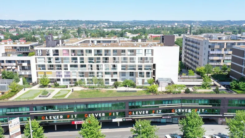 Luftaufnahme eines modernen Gebäudekomplexes mit Geschäftsflächen im Erdgeschoss und Wohnungen darüber, umgeben von viel Grün.