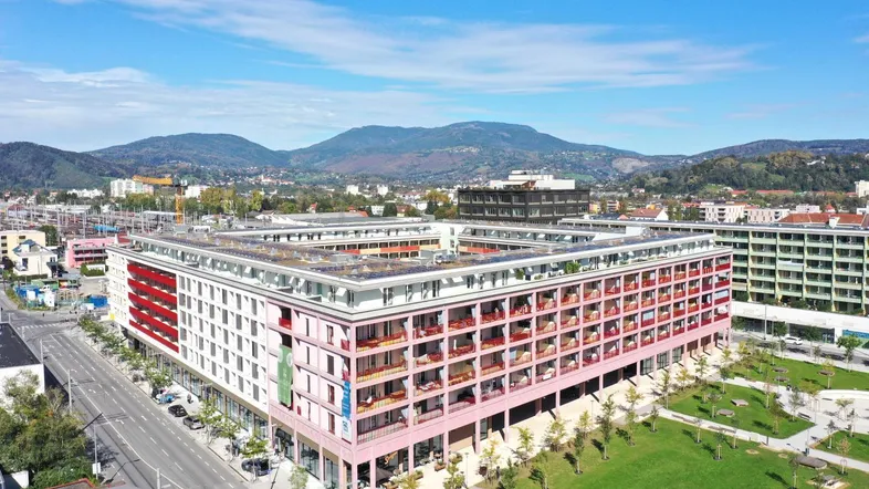 Luftaufnahme eines modernen Gebäudekomplexes mit vielen Balkonen, umgeben von Grünflächen und Bergen im Hintergrund.