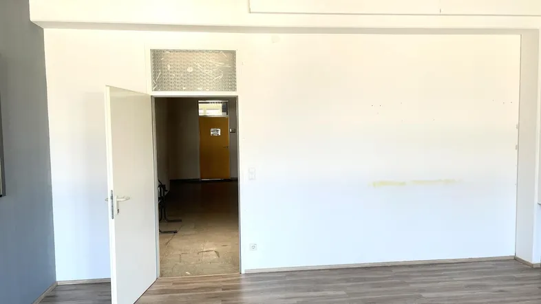 Heller Büroraum mit Laminatboden und einer offenen Tür, die in einen weiteren Bereich führt.
