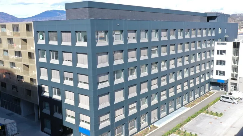 Modernes Bürogebäude mit dunkler Fassade, vielen Fenstern und Parkplätzen in städtischer Umgebung.