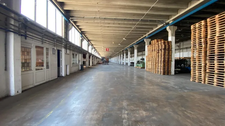 Langer Gang einer Industriehalle mit Betonboden, hohen Decken und seitlichen Fenstern, beleuchtet durch natürliches Licht.