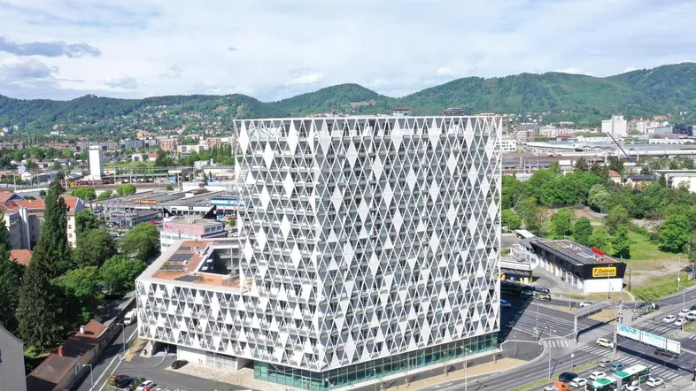 Luftaufnahme eines modernen Gebäudes mit einzigartiger geometrischer Fassade und städtischer Umgebung mit Bergen im Hintergrund.