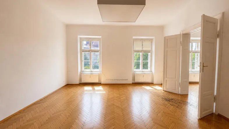 Geräumiges Zimmer mit Fischgrätparkett, hohen Decken und großen Fenstern, die viel Tageslicht hereinlassen.