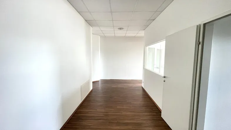 Heller Flur in einem Bürogebäude mit dunklem Laminatboden und weißen Wänden, der zu weiteren Räumen führt.