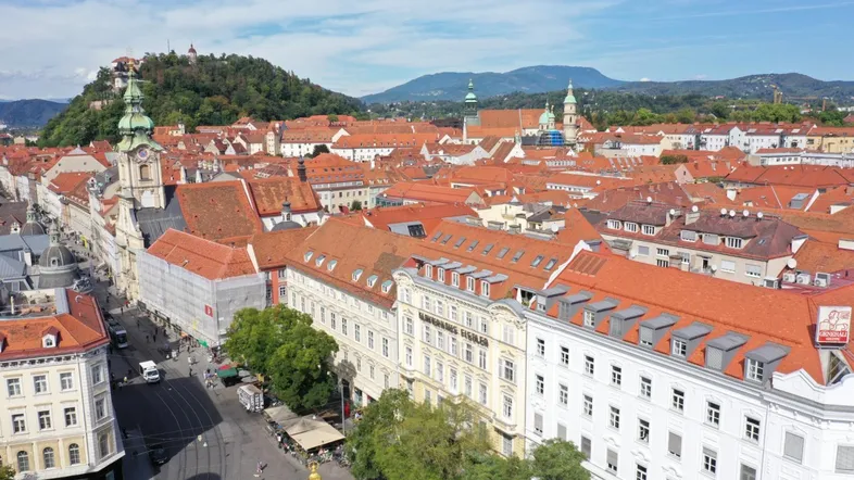 Luftaufnahme einer historischen Stadt mit roten Dächern, einem Schlossberg und Bergen im Hintergrund.