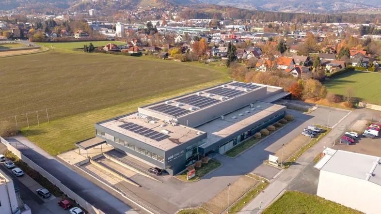 Luftaufnahme eines modernen Geschäftsgebäudes mit Solaranlagen auf dem Dach, umgeben von Feldern und einer bergigen Landschaft.