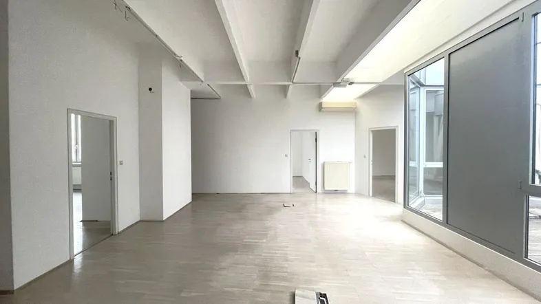 Großer, leerer Innenraum mit hellen Holzböden, weißen Wänden und sichtbaren Deckenbalken, der viel natürliches Licht erhält.