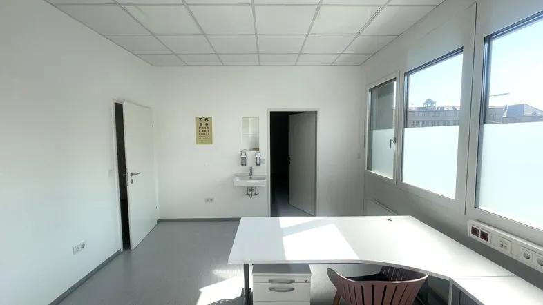 Helles Büro mit großen Fenstern, einem Schreibtisch und einem kleinen Waschbecken, ideal für eine Praxis.