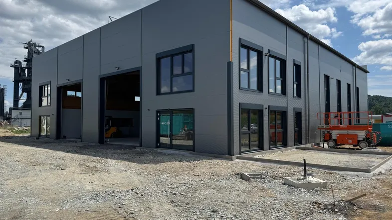 Modernes Gewerbegebäude im Bau mit grauer Fassade und großen Fensterfronten auf einem Schottergrundstück.