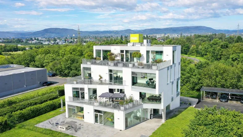 Modernes Gebäude mit mehreren Etagen, großen Glasfronten und Terrassen, umgeben von viel Grün und Bergen.
