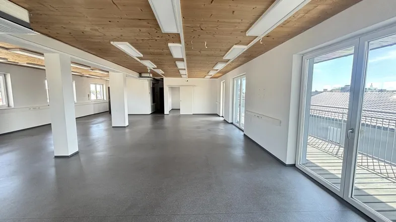 Geräumiger, heller Büroraum mit Holzbalkendecke, Betonboden und Zugang zu einem Balkon.