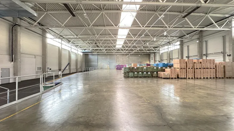 Große, helle Lagerhalle mit hohen Decken, Betonboden und Tageslicht durch Oberlichter.