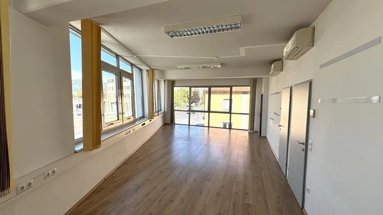 Geräumiger, leerer Büroraum mit großen Fenstern, Holzoptikboden und Klimaanlage, ideal für gewerbliche Nutzung.