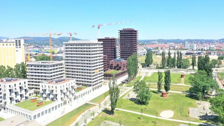 Luftaufnahme einer modernen Stadtentwicklung mit mehreren Gebäuden, Grünflächen und Baukranen im Hintergrund.