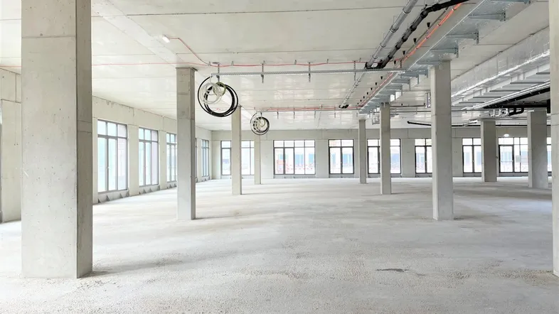 Großer, unfertiger Gewerberaum mit Betonboden, vielen Fenstern und sichtbaren Installationen an der Decke.