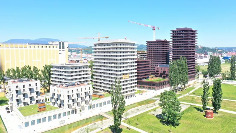 Luftaufnahme eines modernen Wohnkomplexes mit mehreren Gebäuden, Grünflächen und einem Wasserlauf unter blauem Himmel.