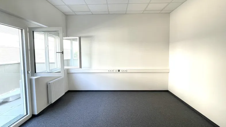 Heller Büroraum mit dunklem Teppichboden, weißen Wänden und Zugang zu einem Balkon.