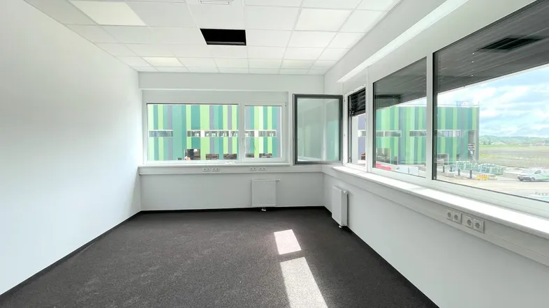 Helles Büro mit Teppichboden und großen Fenstern, die einen Blick auf das Industriegebäude bieten.