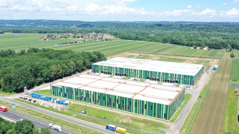 Luftaufnahme von modernen Lagerhallen mit grünen Fassaden, umgeben von Feldern und einer Autobahn.
