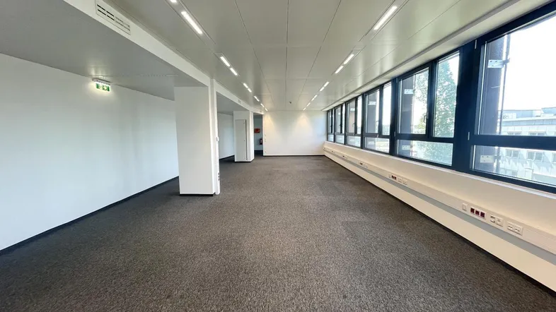 Geräumiger, leerer Büroraum mit Teppichboden, großen Fenstern und moderner Deckenbeleuchtung.