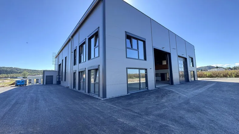 Modernes Industriegebäude mit grauer Fassade, großen Fenstern und viel Freifläche unter blauem Himmel.