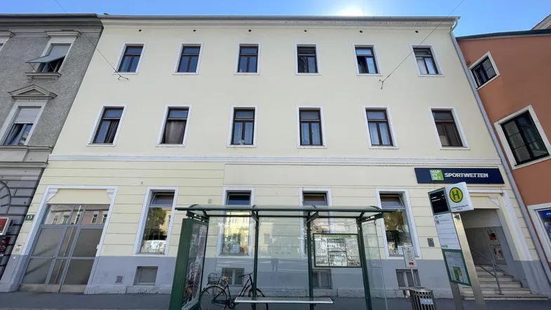 Fassade eines mehrstöckigen Gebäudes mit hellgelber Farbe und vielen Fenstern, davor eine Bushaltestelle.