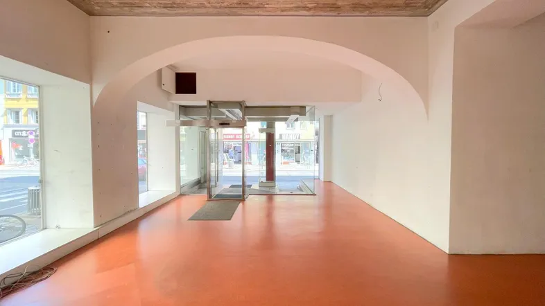 Geräumiger Ladenbereich mit großen Schaufenstern, orangefarbenem Boden und einem markanten Bogen.