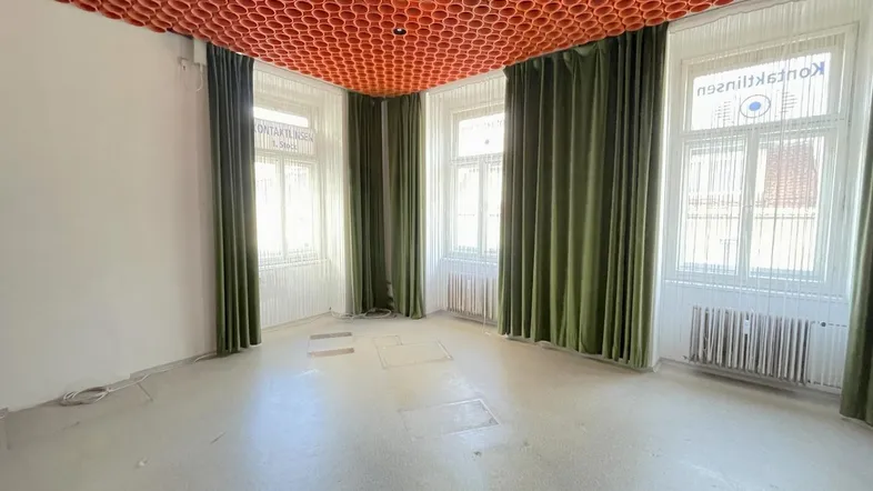 Leerer Raum mit auffälliger orangefarbener Röhrendecke und großen Fenstern mit grünen Vorhängen.