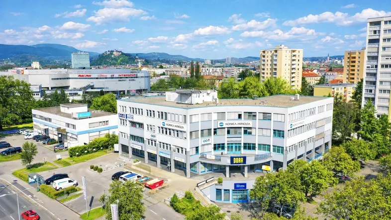 Luftaufnahme eines modernen Geschäftsgebäudes mit Parkplätzen und städtischer Umgebung im Hintergrund.